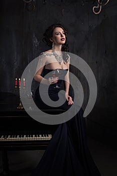 Beautiful young woman in black dress next to a piano with candelabra candles and wine, dark dramatic atmosphere of the castle.