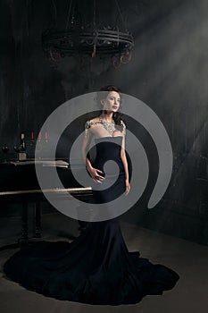 Beautiful young woman in black dress next to a piano with candelabra candles and wine, dark dramatic atmosphere of the castle.