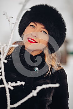 A beautiful young woman in a black fur hat and fur coat, portrait. A cute Russian girl smiles and looks into the frame