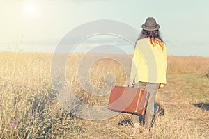 Beautiful young woman with black hat carries brown vintage suitcase in the field road during summer sunset. Back view. Toned image