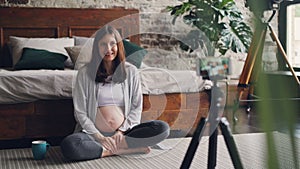 Beautiful young woman blogger is recording video about pregnancy for her vlog sitting at home on the floor, talking and