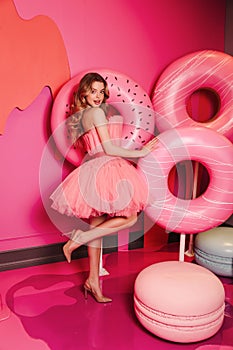 Beautiful young woman with blond hair in elegant dress posing in pink room with decoration of desserts