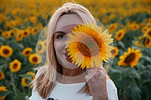 A beautiful young woman with blond hair holds a large sunflower flower in her hands and covers one eye with it. Against