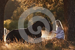 Beautiful young woman with blond hair sitting in front of lying horse and feed it from hands in sunset sunlight in autumn