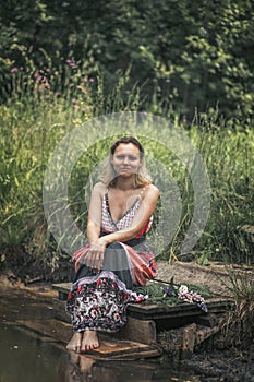 A beautiful young woman with blond hair in a sundress on the shore of a lake with a bouquet of wildflowers. National Russian