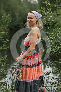 A beautiful young woman with blond hair in a sundress on the shore of a lake with a bouquet of wildflowers. National Russian