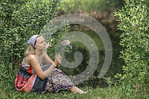 A beautiful young woman with blond hair in a sundress on the shore of a lake with a bouquet of wildflowers. National Russian