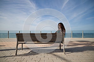 beautiful young woman, blonde with curly hair and blue eyes is sitting on a bench looking out to sea. The woman has the Atlantic