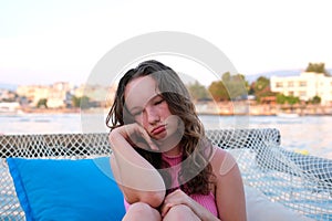 beautiful young woman, blonde with curly hair and blue eyes is sitting on a bench looking out to sea. The woman has the