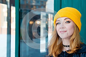 Beautiful young woman with blonde hair and blue eyes in a yellow knitting hat on a background of green wall.