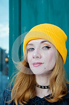 Beautiful young woman with blonde hair and blue eyes in a yellow knitting hat on a background of green wall.