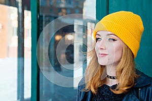 Beautiful young woman with blonde hair and blue eyes in a yellow knitting hat on a background of green wall.