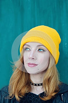 Beautiful young woman with blonde hair and blue eyes in a yellow knitting hat on a background of green wall.