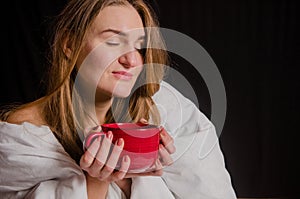 Beautiful young woman with blonde hair, sits in the morning wrapped in a white blanket, drinks tea from red cup