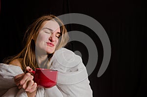 Beautiful young woman with blonde hair, sits in the morning wrapped in a white blanket, drinks tea from red cup