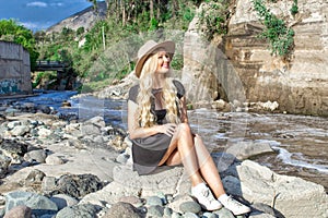 A beautiful young woman blonde with long hair in a hat sitting on a rocky shore by the river. Around the mountains and rocky terra