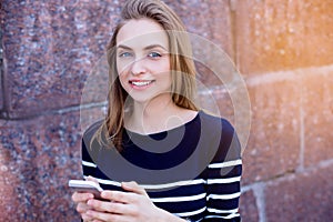 Beautiful young woman, blonde, making reminders outdoors using smartphone and fast 4G internet connection while standing against s