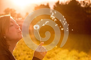 Beautiful young woman is blowing dandelion, evening sun