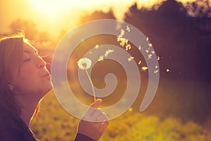 Beautiful young woman is blowing dandelion, evening sun