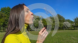 Beautiful young woman blowing a dandelion. Girl with dandelion in hand. Allergic to pollen of flowers. Spring allergy