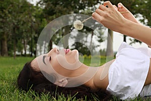 Beautiful young woman blowing dandelion while lying on green grass in park. Allergy free concept