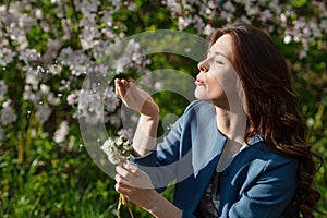 Beautiful young woman blowing dandelion in spring garden. Enjoy Nature. Healthy smiling girl outdoor. Allergy free concept.