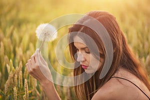 Beautiful young woman blows dandelion in a wheat field in the summer sunset. Beauty and summer concept