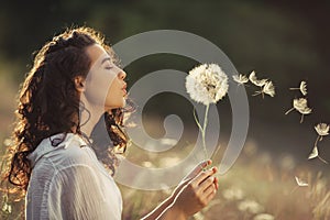 Beautiful young woman blows dandelion in a wheat field in the summer sunset. Beauty and summer concept