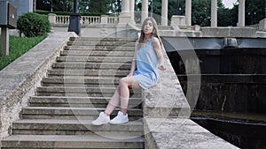 Beautiful young woman in blue dress sits on staircase near cascading fountain