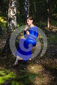 Beautiful, young woman in a blue dress is sitting near a tree over a cliff, against a forest background.