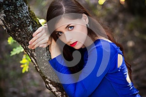 Beautiful, young woman in a blue dress is sitting near a tree over a cliff, against a forest background.