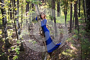 Beautiful, young woman in a blue dress stands near a tree over a cliff, against a forest background.