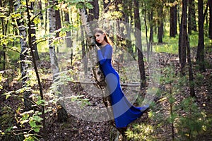 Beautiful, young woman in a blue dress stands near a tree over a cliff, against a forest background.