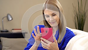 Beautiful young woman in blue t-shirt with smartphone sitting on sofa. Browsing Internet smiling