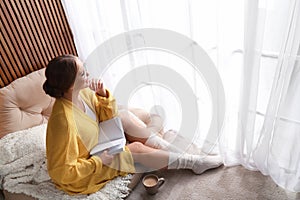 Beautiful young woman with book and cup of coffee near window at home. Winter atmosphere