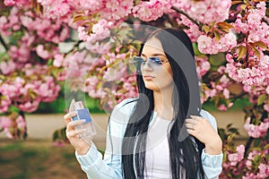 Beautiful young woman with bottle of perfume. Spring day. Spring pink sakura blossom. Fashionable girl in trendy glasses. Women