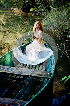 Beautiful young woman, bride, with red hair, Lady of Shalott, sits eagerly abandoned with a bouquet of flowers on a boat and waits