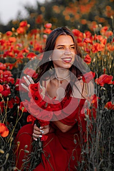 Beautiful young woman with brown hair in red long dress in field with poppies in evening at sunset and holds poppy in hand, Summer