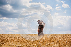Beautiful young woman with brown hear wearing rose dress and hat enjoying outdoors looking to the horizont on perfect wheat field