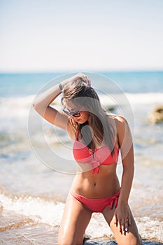 Beautiful young woman with brunette hair in pink swimsuit is posing on the beach near rocks on the background of sea