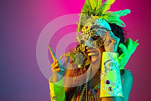 Beautiful young woman in carnival and masquerade costume on gradient studio background in neon light