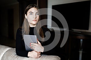 Beautiful young woman in casual clothes is using a digital tablet and smiling while sitting on couch at home