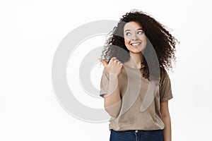 Beautiful young woman in casual outfit pointing finger left, looking aside at banner with happy white smile, showing