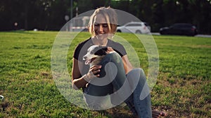 Beautiful young woman in casual sitting on grass with her dog pet and smiling. Owner with lovely little dog outdoors