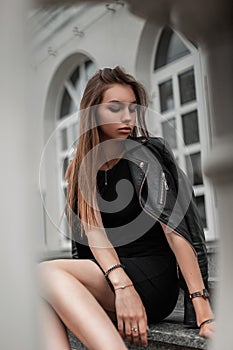 Beautiful young woman with chic brown long hair in a black stylish dress in a vintage black leather jacket posing near a white