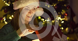 Beautiful young woman in a Christmas hat opening Christmas gift box with excited surprised face feeling happy on New Year`s Eve