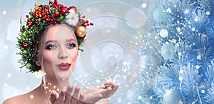 Beautiful young woman with Christmas wreath blowing magical snowy dust on blurred background