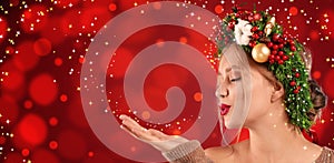 Beautiful young woman with Christmas wreath blowing magical snowy dust on red background