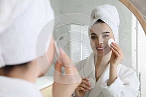 Beautiful young woman cleaning her face with cotton disk near mirror in bathroom