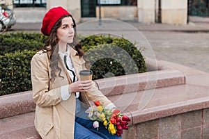 Beautiful young woman with coffee to go and spring tulips flowers bouquet at city street. Happy girl walking outdoors. Spring